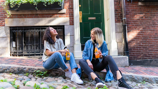Two women sitting on a cobblestone street, laughing and holding orange soda bottles.