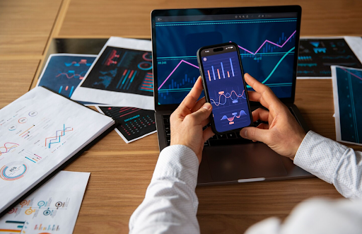 A person holding a smartphone displaying financial data charts over a laptop and desk covered in printed analytical reports and graphs